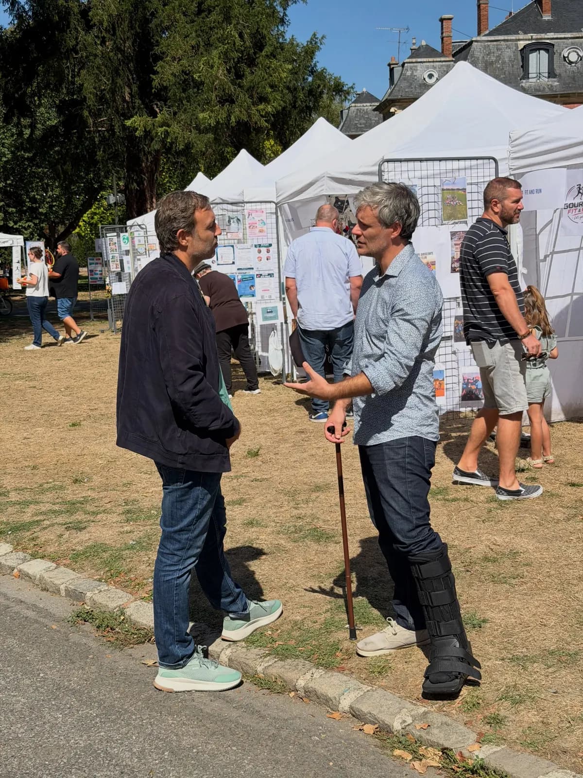 Deux hommes discutent debout, devant l’alignement des tentes blanches des stands installés sur une pelouse.