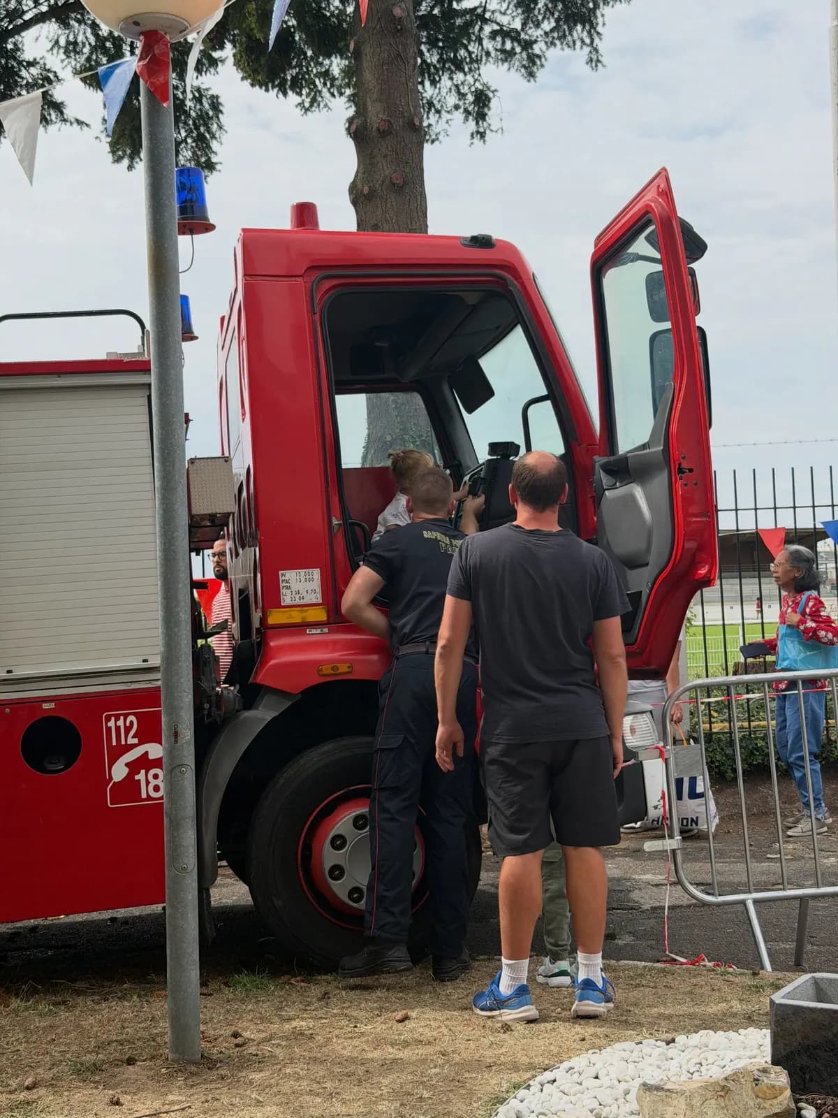 Un camion de pompiers rouge, portière ouverte, devant lequel des visiteurs attendent sous des guirlandes de fanions.