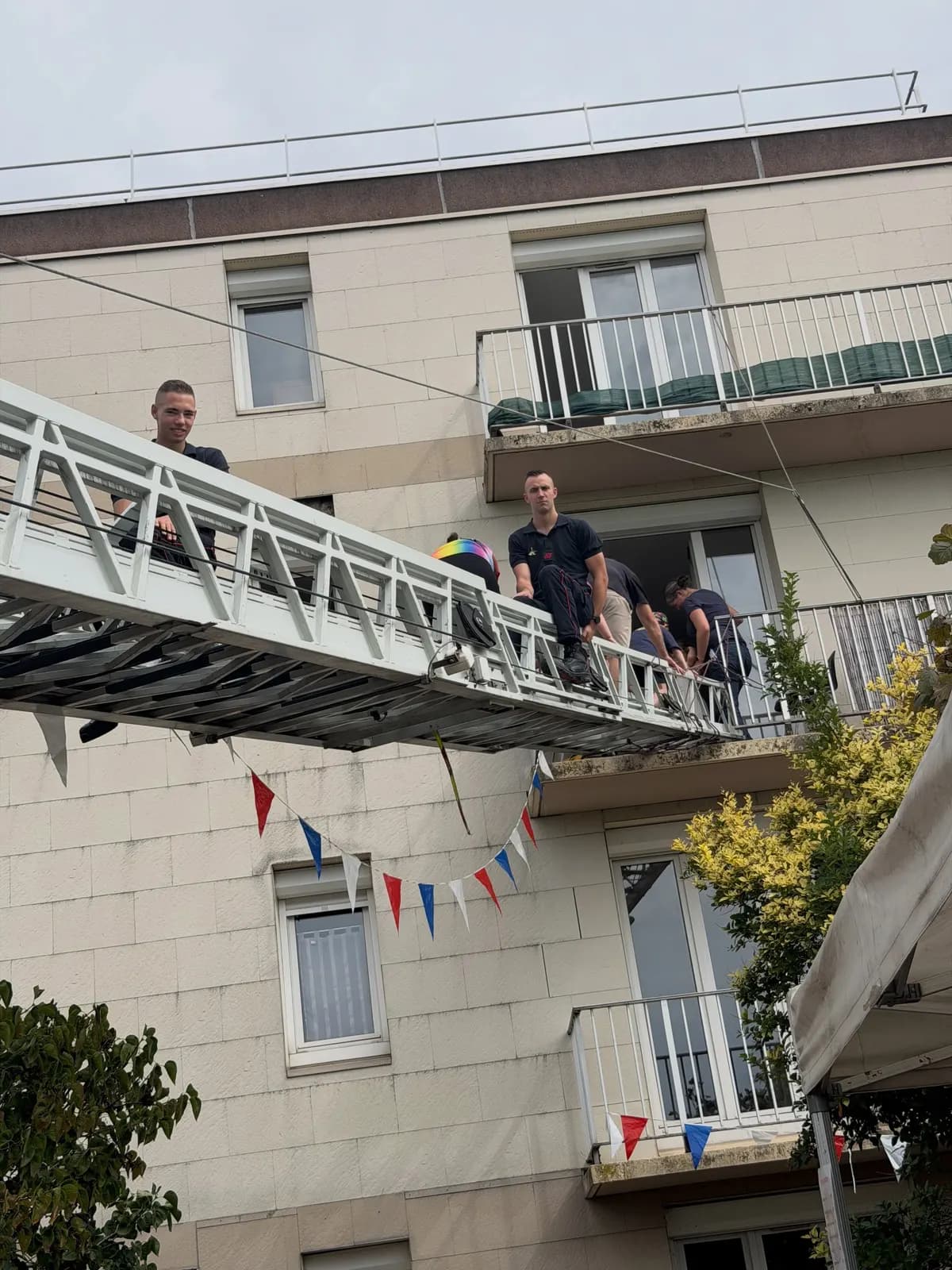 Une grande échelle déployée le long d’une façade d’immeuble, avec des pompiers et des visiteurs sur les échelons.