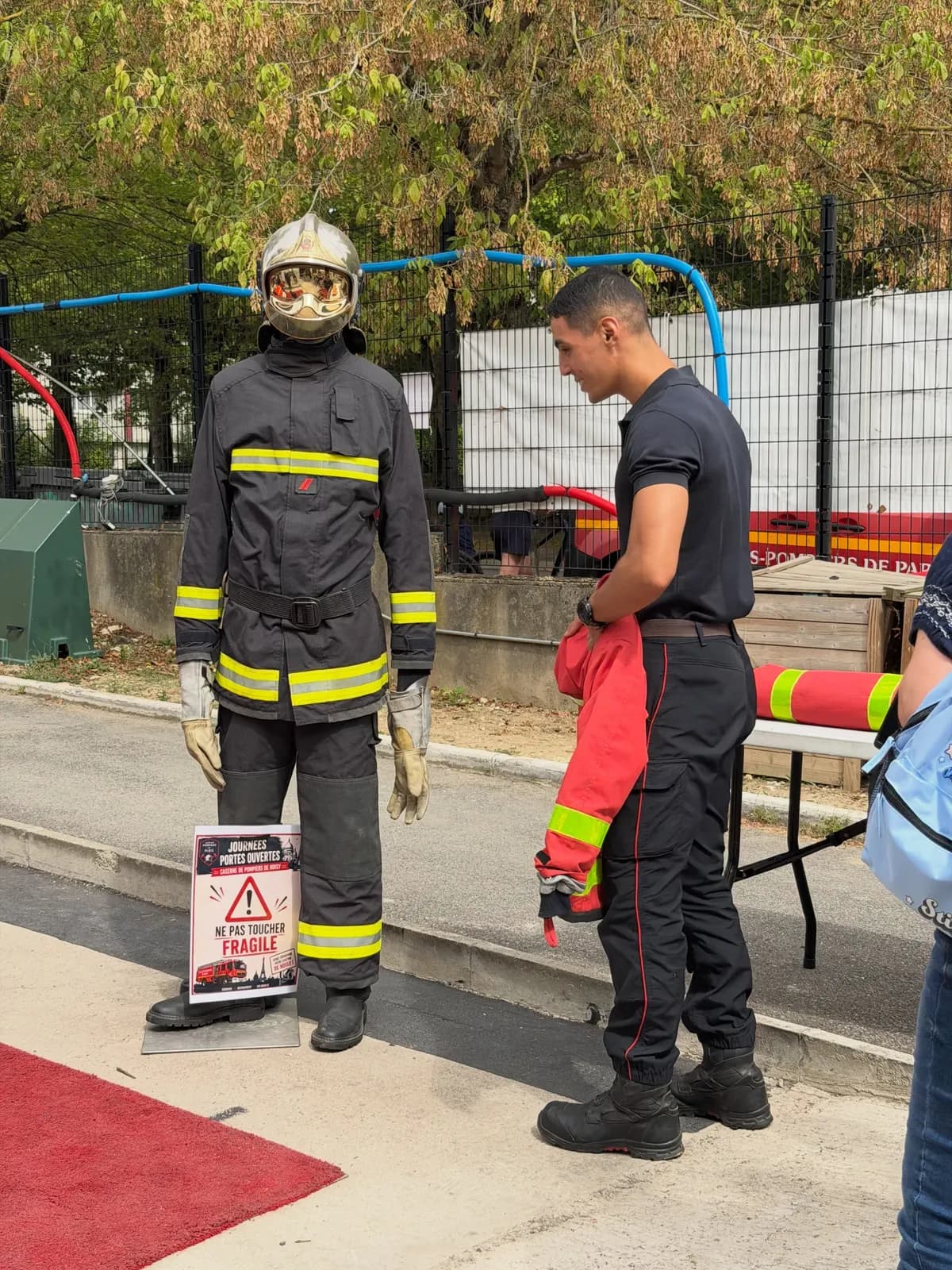 Tenue de sapeur-pompier sur un mannequin, panneau « Ne pas toucher, fragile », à côté d’un pompier tenant une veste.