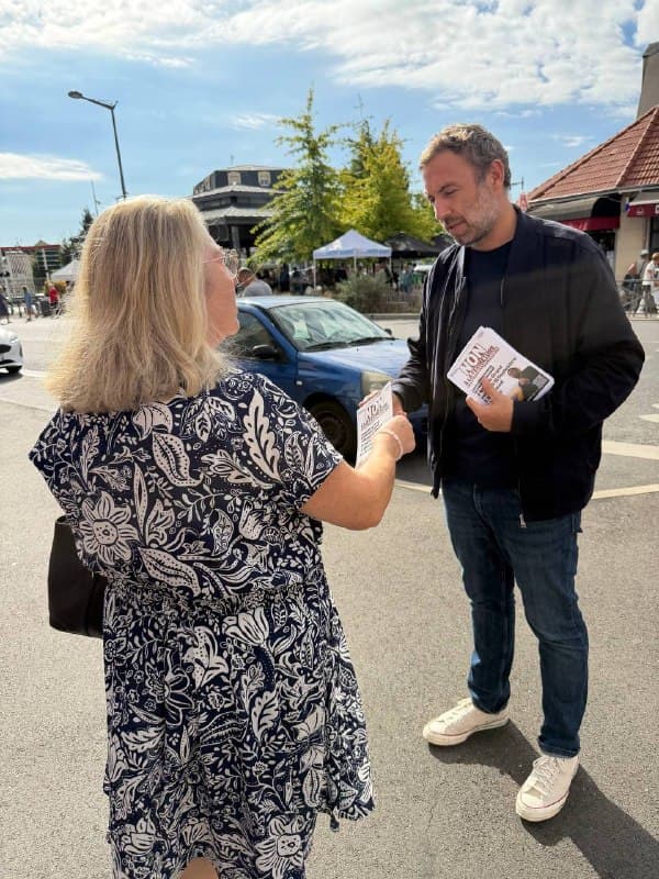 Un homme en veste sombre tend un tract à une passante en robe fleurie, sur une place.