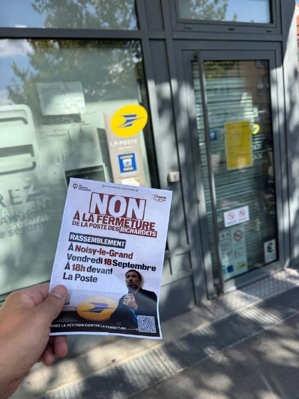 Une main tient un tract « Non à la fermeture de la Poste des Richardets » devant la porte d’un bureau de poste.