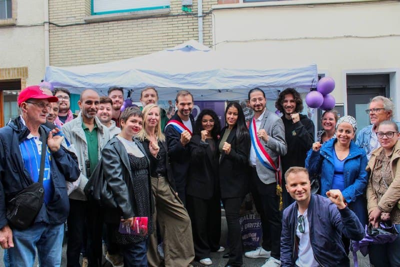 Une vingtaine de militants posent devant un stand sous barnum orné de ballons violets, dans une rue ; plusieurs lèvent le poing, trois portent une écharpe tricolore.