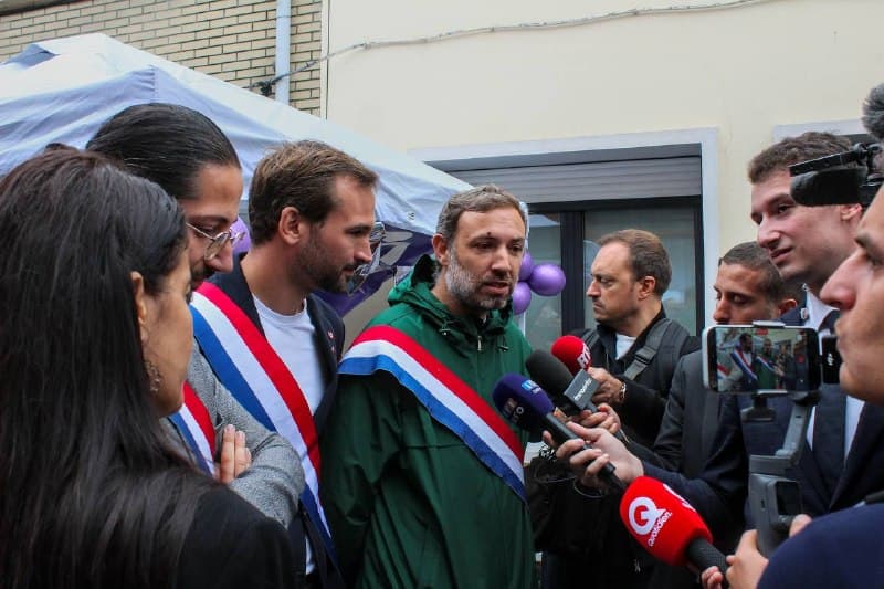 Des élus en écharpe tricolore répondent aux micros de plusieurs journalistes devant un stand.