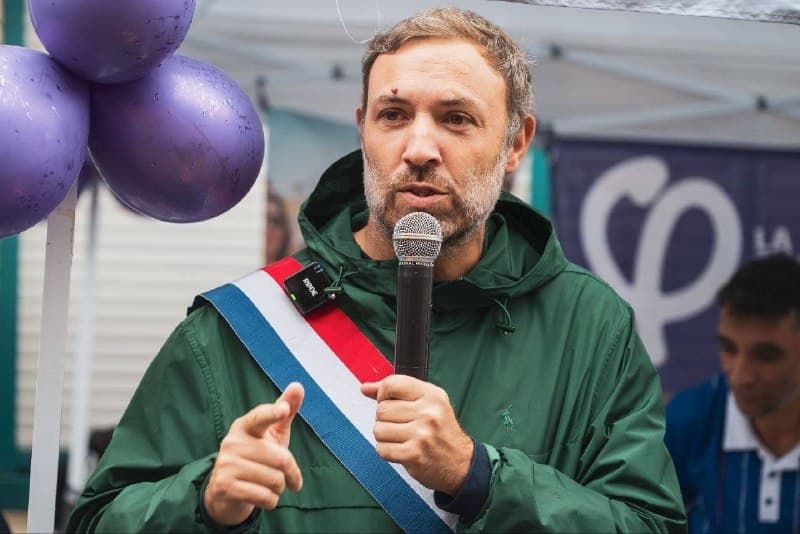 Un élu en veste verte et écharpe tricolore parle au micro devant des ballons violets et une banderole au logo de La France insoumise.