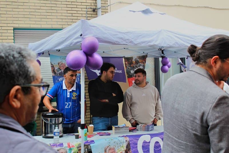 Des militants tiennent un stand sous barnum, avec ballons violets, tracts et thermos, face à des passants.