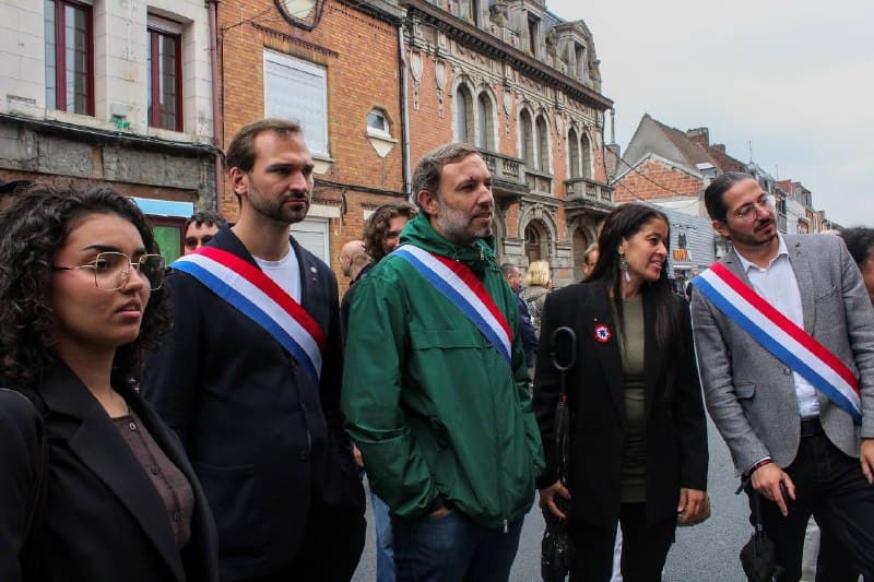 Cinq personnes, dont trois élus en écharpe tricolore, se tiennent dans une rue bordée de maisons de brique.