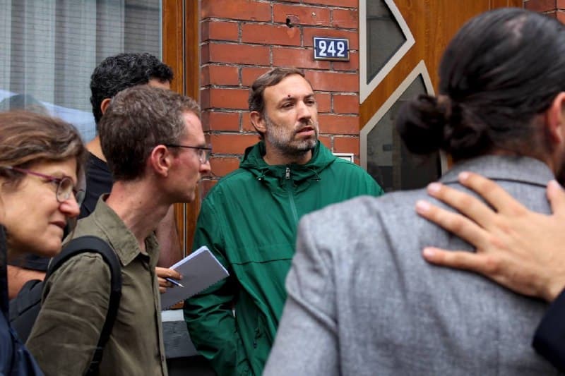 Un élu en veste verte discute avec des passants devant une porte de brique numérotée 242.