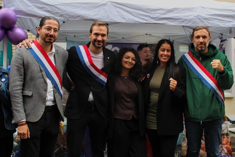 Cinq personnes posent devant un stand, dont trois élus en écharpe tricolore ; deux lèvent le poing.