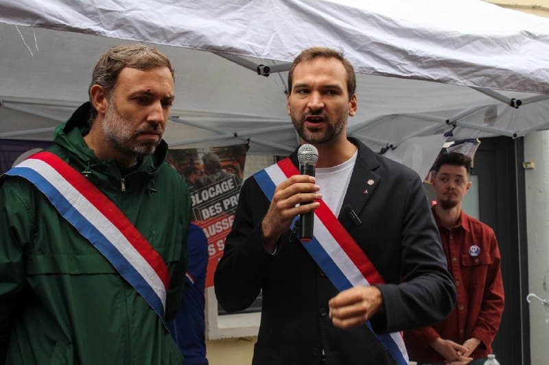 Un élu en écharpe tricolore parle au micro sous un barnum, à côté d’un autre élu en veste verte.