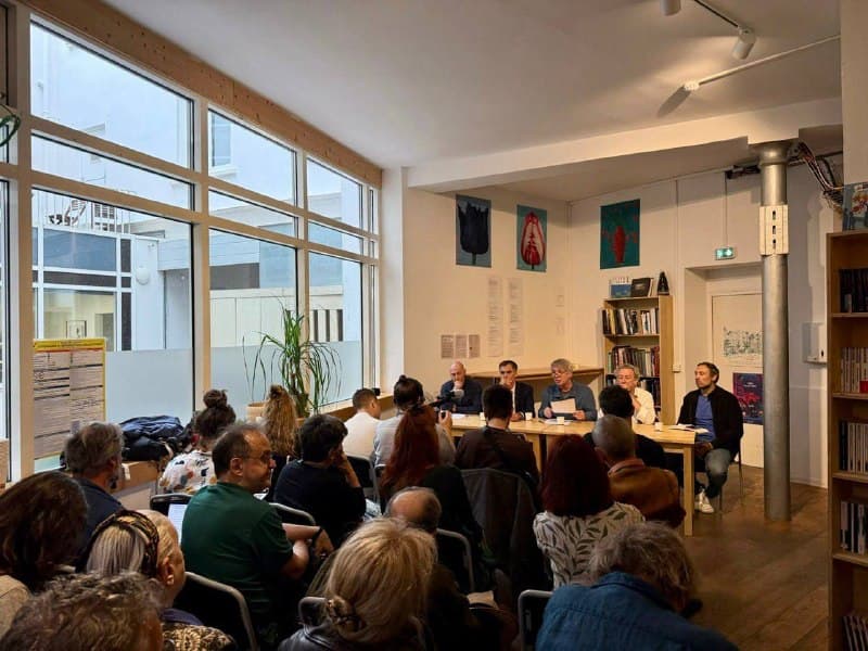 La même réunion vue de plus loin : le public, assis en rangs serrés, écoute les intervenants installés au fond de la salle.