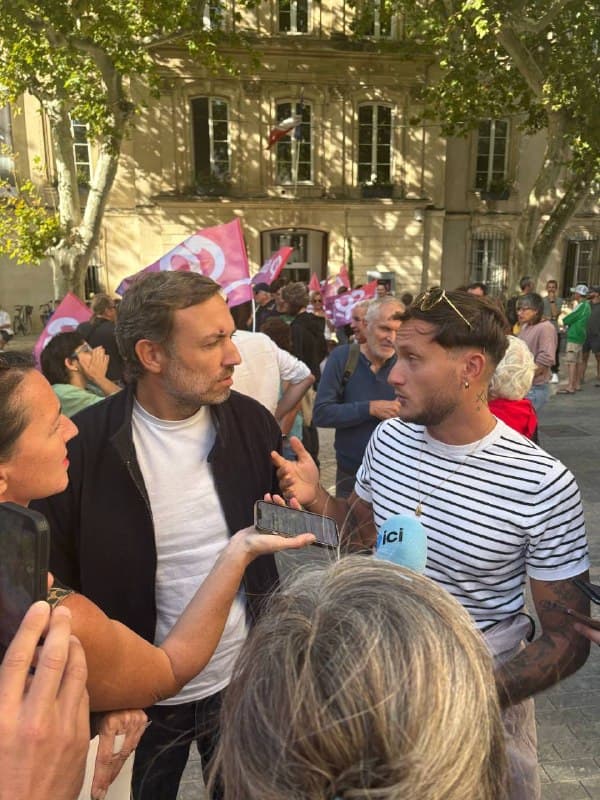 Dans la foule d’un rassemblement, sous des drapeaux, un homme en veste sombre échange avec un jeune homme en marinière ; un micro de la radio ici est tendu vers eux.