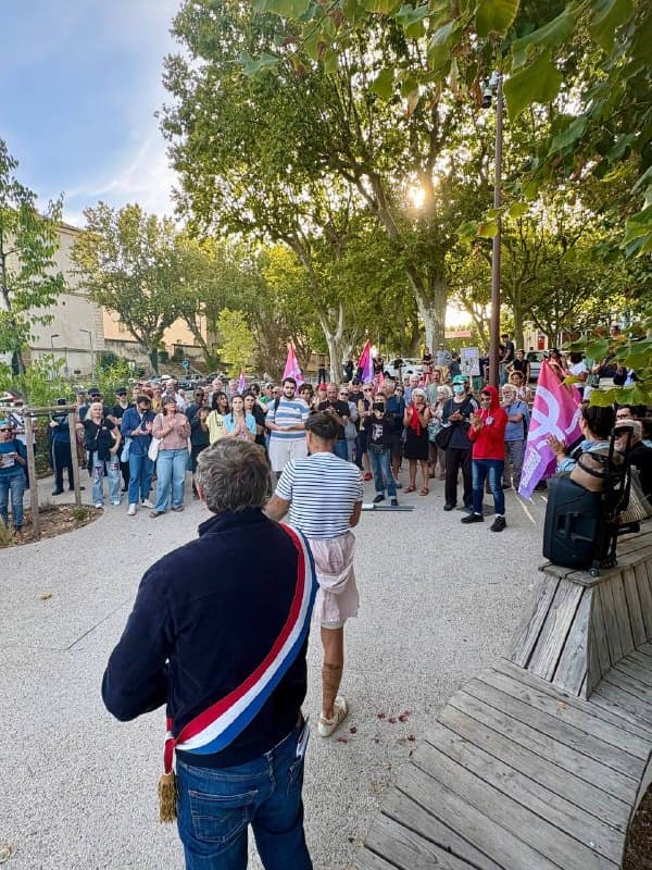 Vu de dos, un élu en écharpe tricolore fait face à une foule rassemblée sur une place ombragée, où flottent des drapeaux.