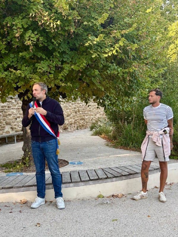 Deux hommes debout sur une place arborée, l’un en écharpe tricolore, l’autre en marinière et short.