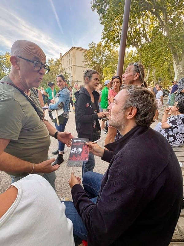 Assis sur un banc, un homme tient un livre face à un groupe de personnes debout, sur une place où se tient un rassemblement.
