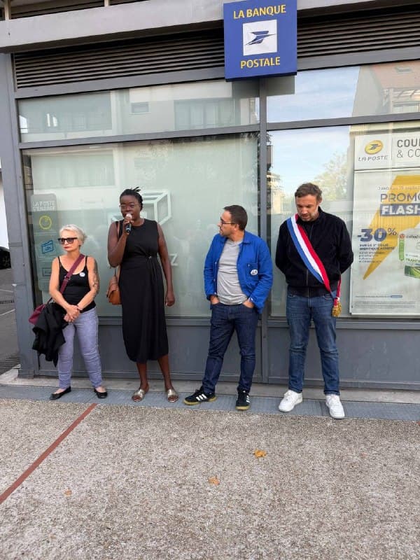 Quatre personnes devant La Banque postale, dont une femme au micro et un homme en écharpe tricolore.