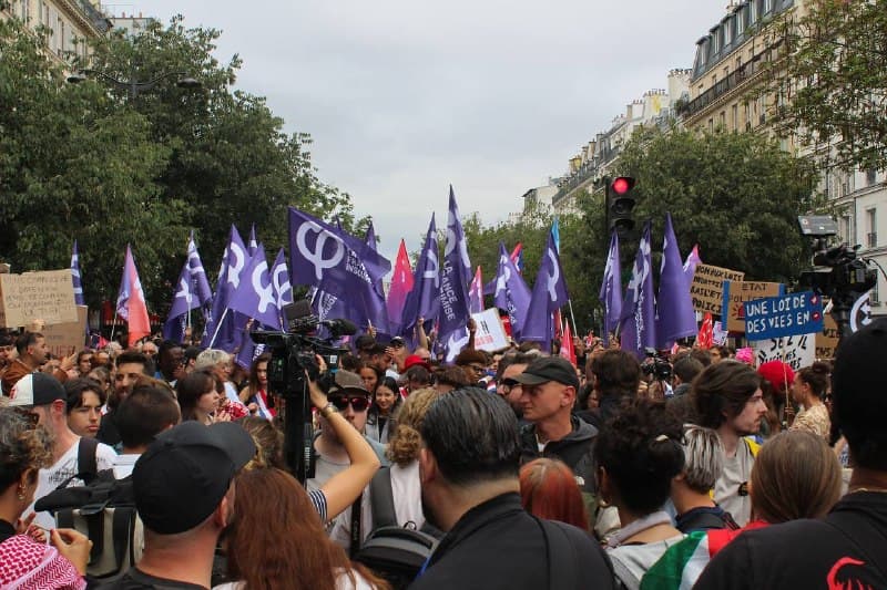Le cortège sur un boulevard parisien, drapeaux violets levés, pancartes et caméras au-dessus de la foule.