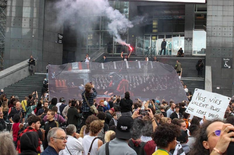 Grande toile déployée devant des marches, fumigène rouge, pancarte « Nos vies valent plus que leurs balles ».
