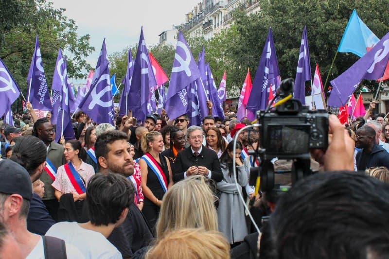 Élus en écharpe tricolore et un homme aux cheveux blancs en costume, filmés au milieu des drapeaux violets.
