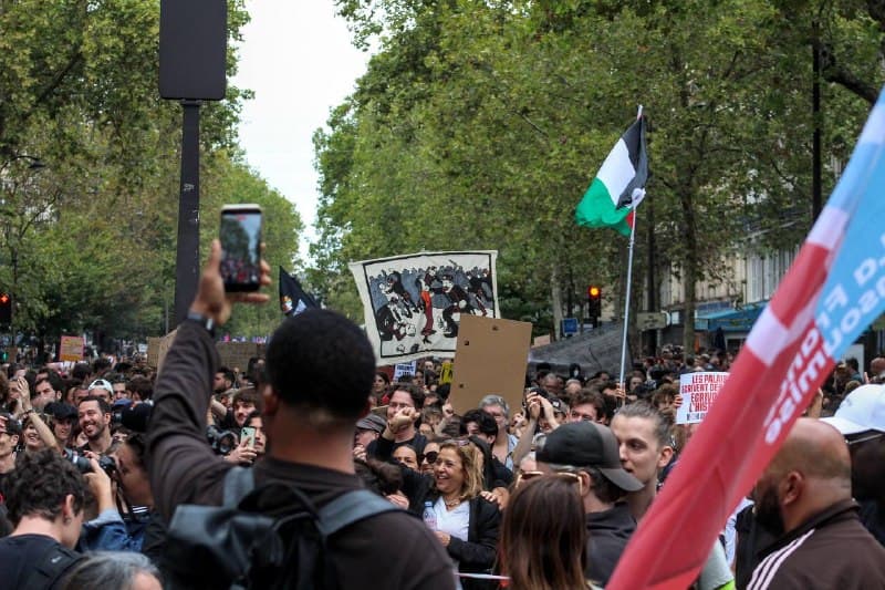 La foule du cortège sous les arbres, un drapeau palestinien et une toile levés, un téléphone tendu au premier plan.