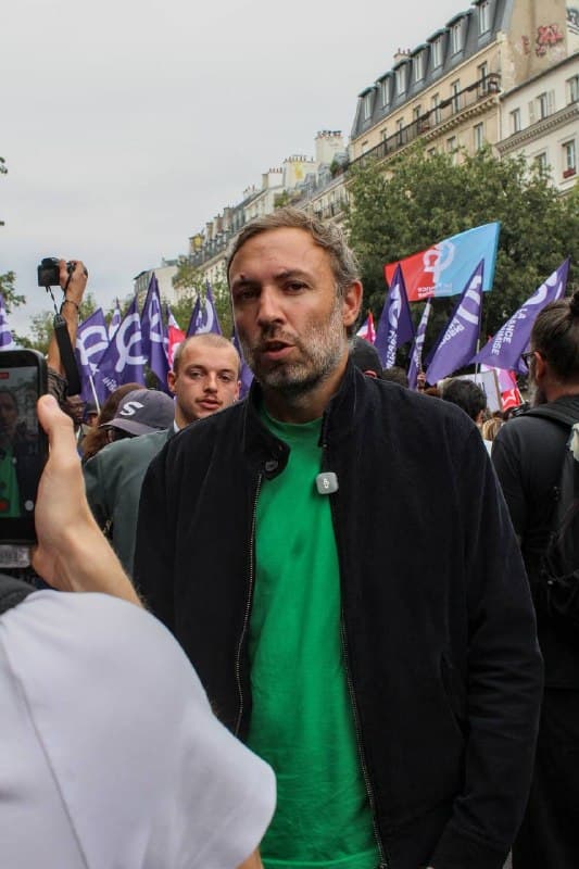 Portrait d’un homme au tee-shirt vert et veste noire dans le cortège, drapeaux violets et roses derrière lui.