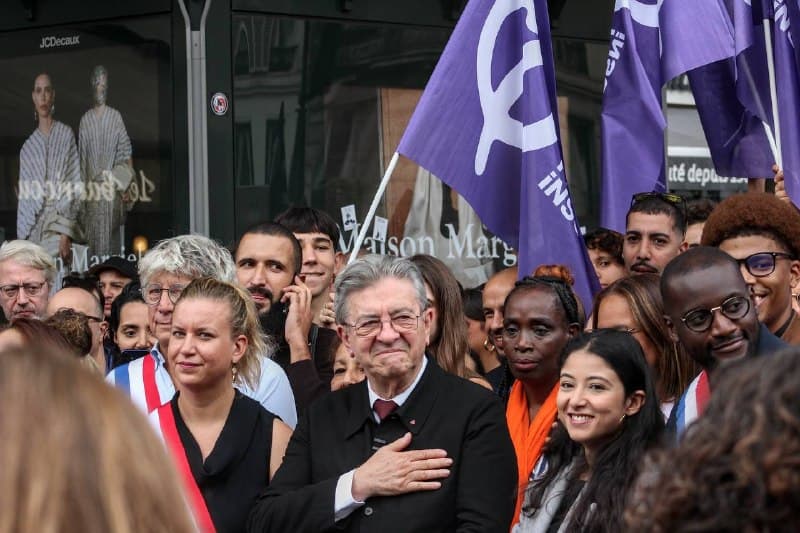 Un homme aux cheveux blancs, la main sur le cœur, entouré d’élues en écharpe tricolore et de drapeaux violets.