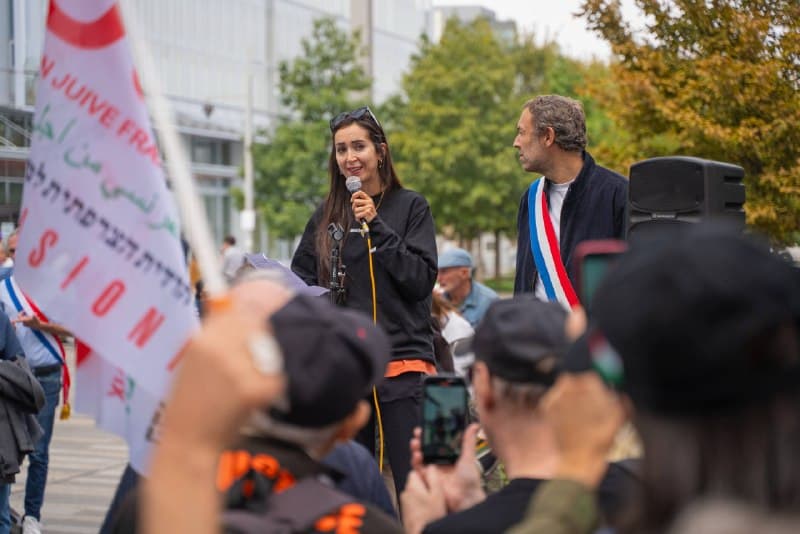 Une oratrice au micro pendant le rassemblement, Thomas Portes en écharpe tricolore derrière elle.