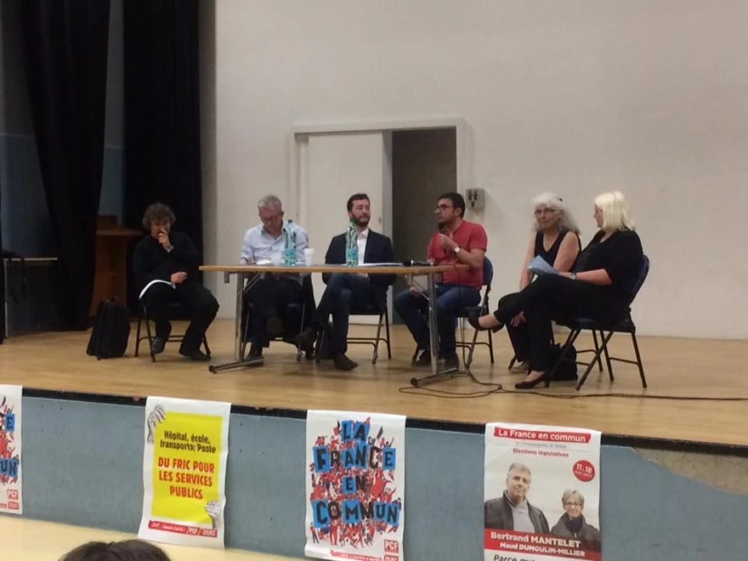 Six personnes assises à une table sur scène lors d'une réunion publique, devant des affiches syndicales et politiques