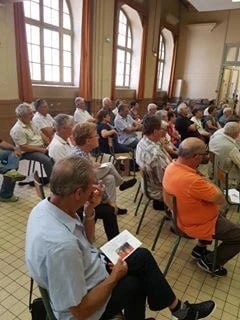 Photographie du public assis sur des chaises dans une salle aux hautes fenêtres, vu de dos.