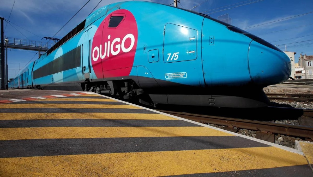 Train à grande vitesse bleu et rose « Ouigo » à quai, vu en contre-plongée depuis un marquage jaune et noir.