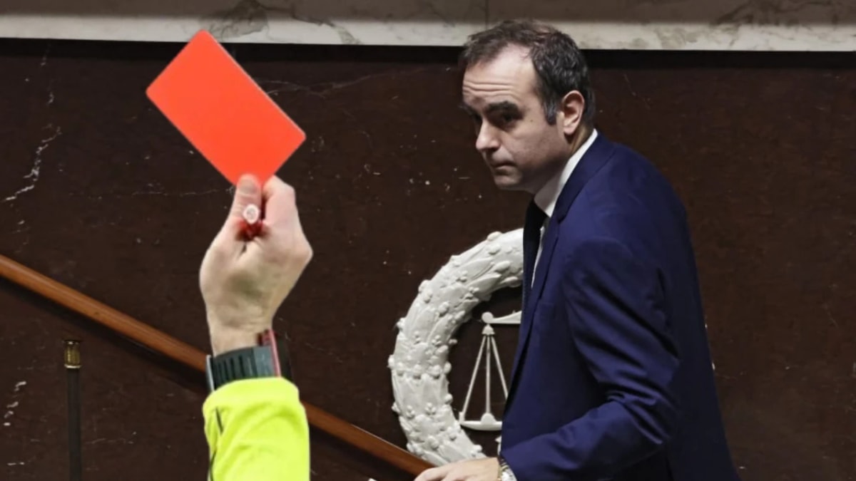 Photographie d’une main tenant un carton rouge au premier plan, face à un homme en costume qui se retourne dans l’hémicycle.