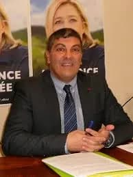 Homme souriant en costume, assis à une table couverte de documents, devant des affiches au portrait d'une femme