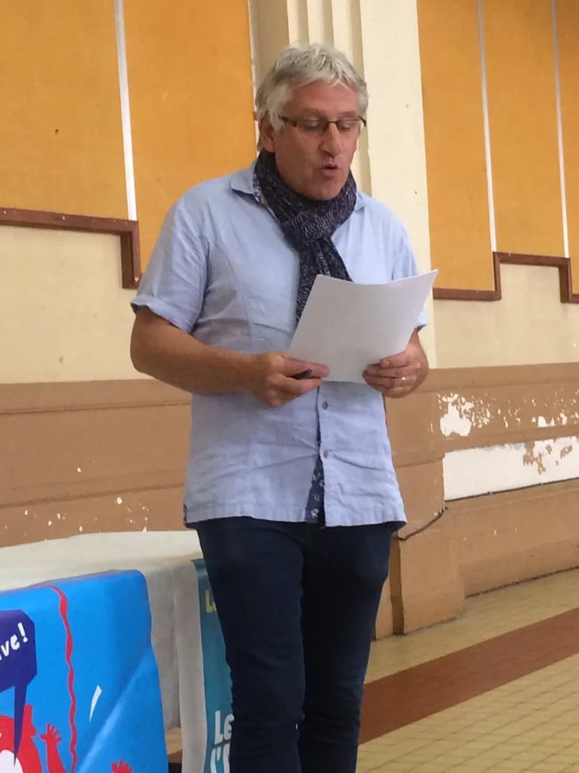 Photographie d'un homme aux cheveux gris, en chemise bleue et écharpe, lisant un texte sur une feuille debout dans une salle.