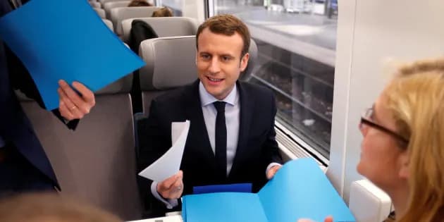 Homme souriant en costume assis dans un train, tenant des feuilles, face à une femme et à des chemises cartonnées bleues