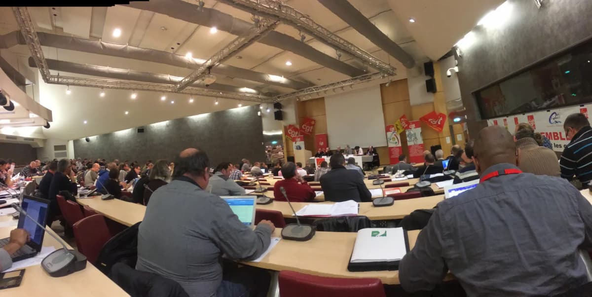 Salle de conférence en gradins, participants attablés devant des ordinateurs portables, drapeaux au fond.