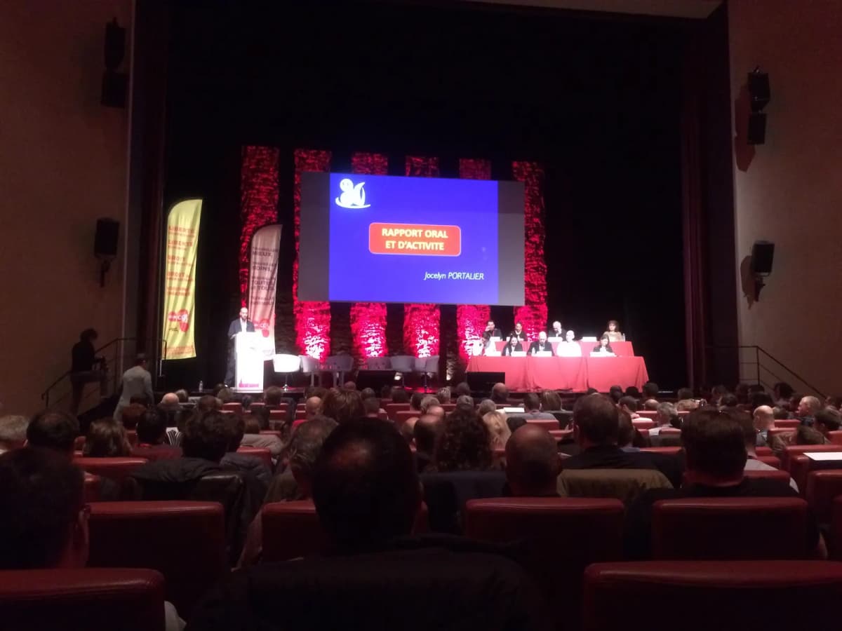 Salle de congrès vue depuis le fond, tribune éclairée et écran affichant un rapport d’activité.