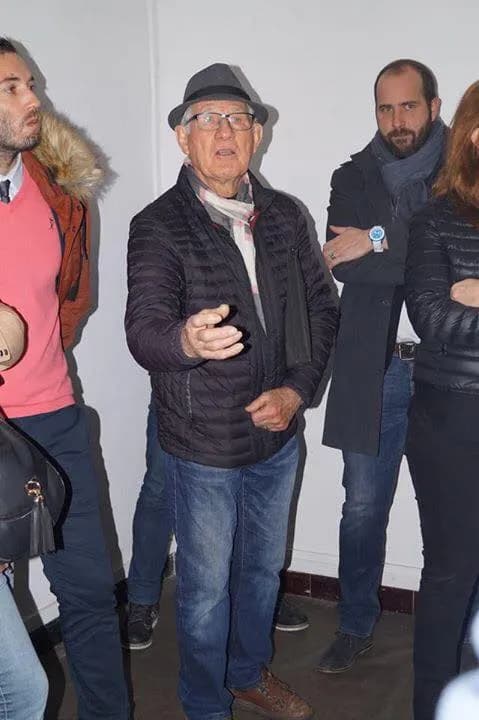 Un homme âgé coiffé d’un chapeau parle en faisant un geste de la main, entouré de plusieurs personnes.