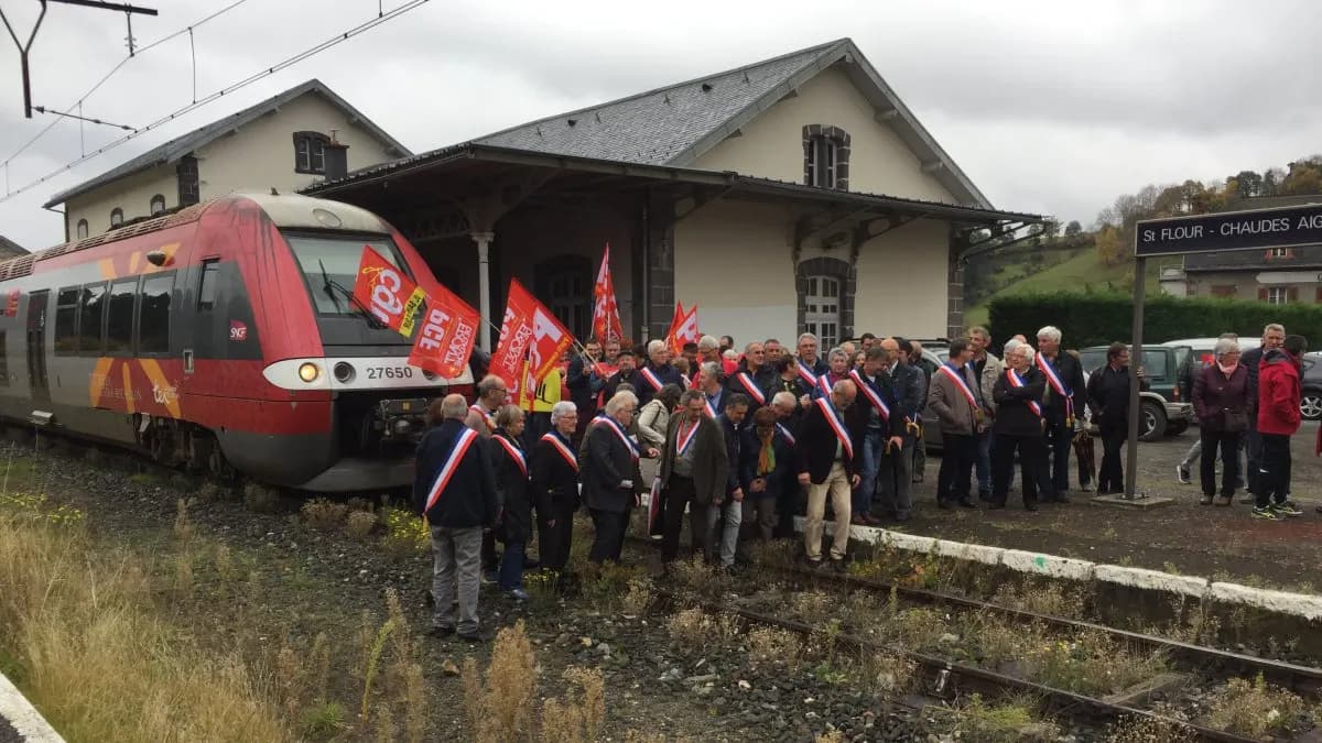Rassemblement dans une petite gare devant un autorail, plusieurs personnes en écharpe tricolore, drapeaux syndicaux