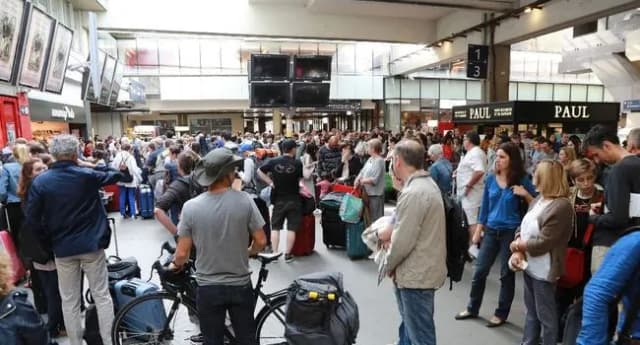 Hall de gare bondé de voyageurs avec leurs bagages devant les commerces.