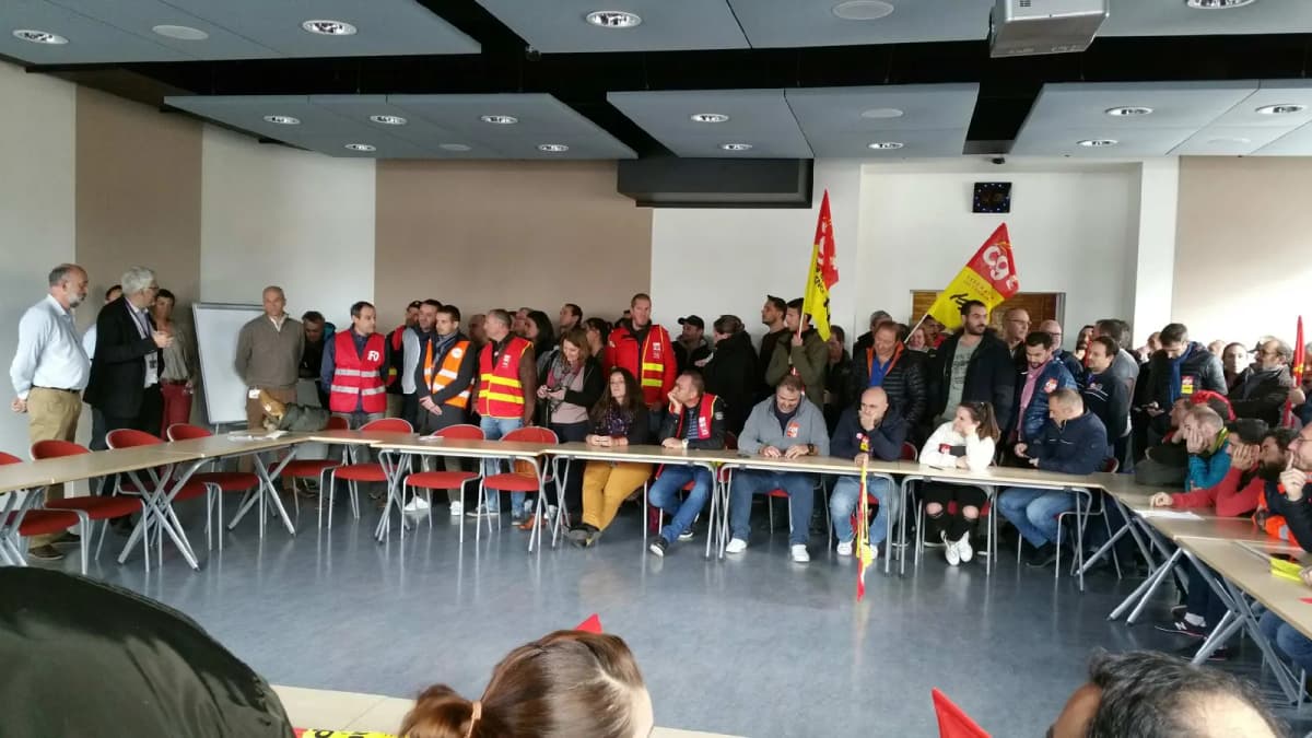 Assemblée syndicale dans une salle, participants en gilets de haute visibilité et drapeaux CGT au fond.