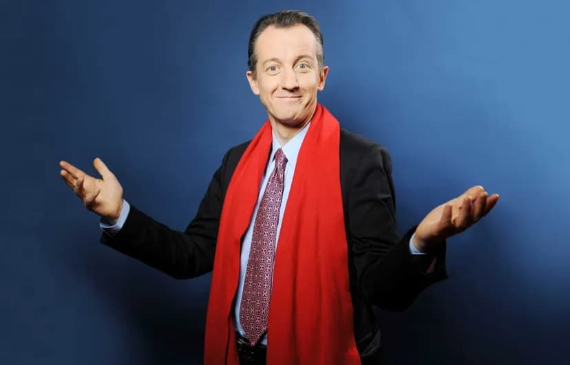 Photographie en studio d'un homme en costume et écharpe rouge, souriant, les bras ouverts paumes vers le haut, sur fond bleu.