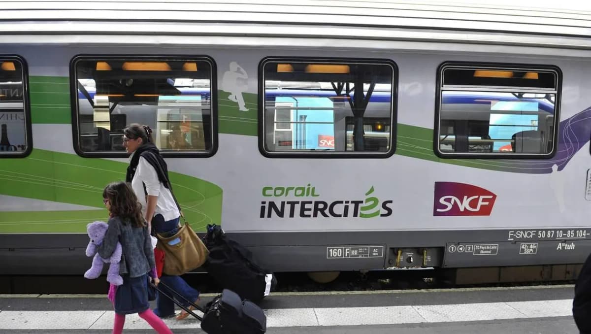 Une adulte et une enfant marchent avec leurs bagages le long d’une voiture Corail Intercités à quai.