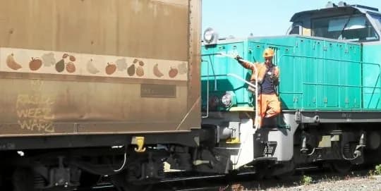 Wagon orné de fruits peints attelé à une locomotive verte, un agent en tenue orange sur le marchepied.
