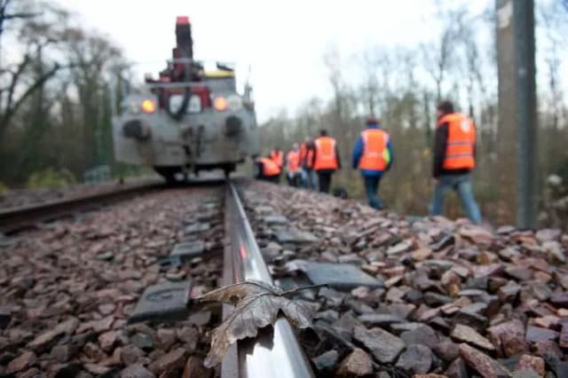 Voie ferrée, une feuille morte sur le rail au premier plan, des agents en gilet orange et un engin de chantier au loin.