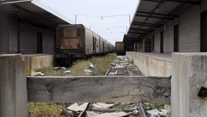 Voie de débord envahie d’herbe entre deux quais de marchandises désaffectés, un train à l’arrêt au fond.