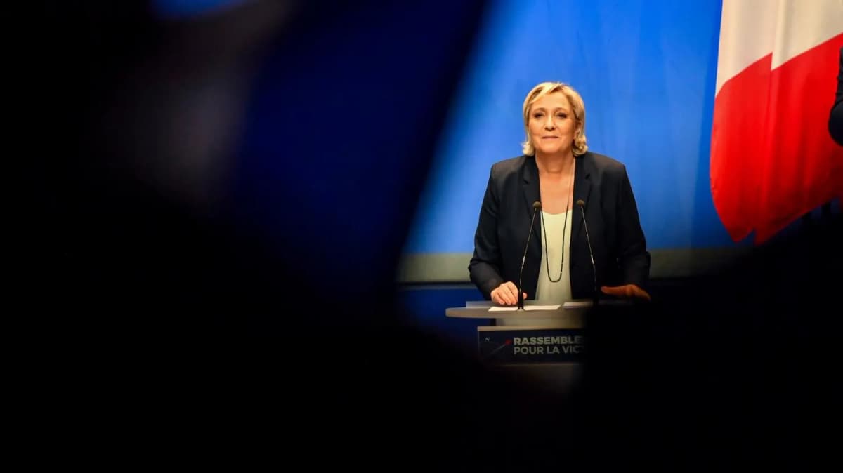 Une femme debout à un pupitre portant « Rassemble… pour la vict… », devant un drapeau français, entre des silhouettes floues.