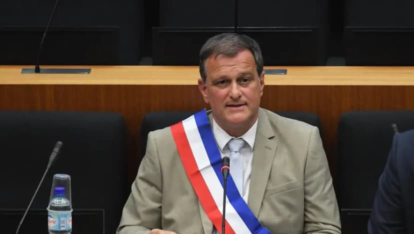 Un homme en costume beige portant l’écharpe tricolore, assis devant un micro dans une salle de conseil.