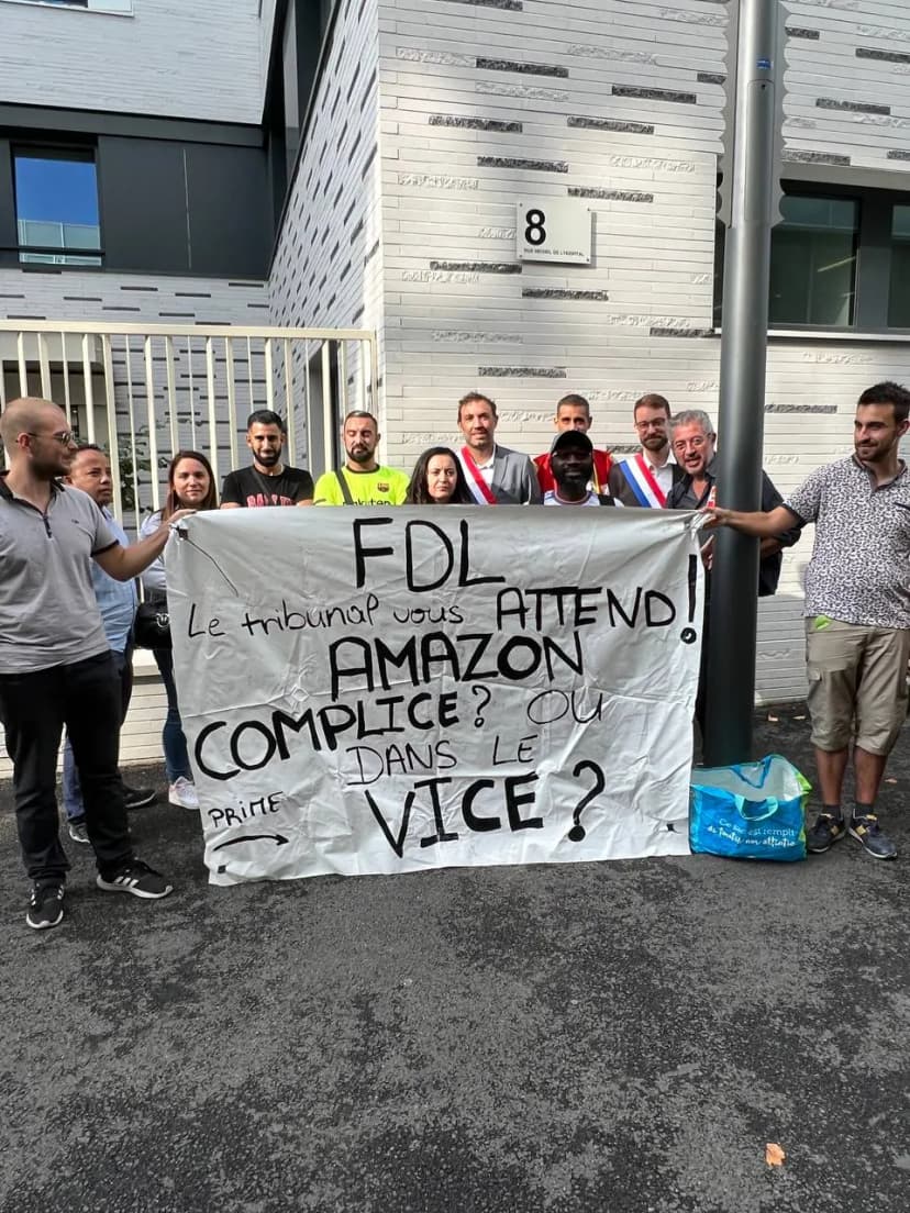 Un groupe déploie une banderole manuscrite devant l’entrée d’un bâtiment, plusieurs personnes portant l’écharpe tricolore.