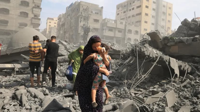Une femme porte un enfant dans ses bras au milieu des décombres d’immeubles détruits, d’autres personnes marchent derrière.