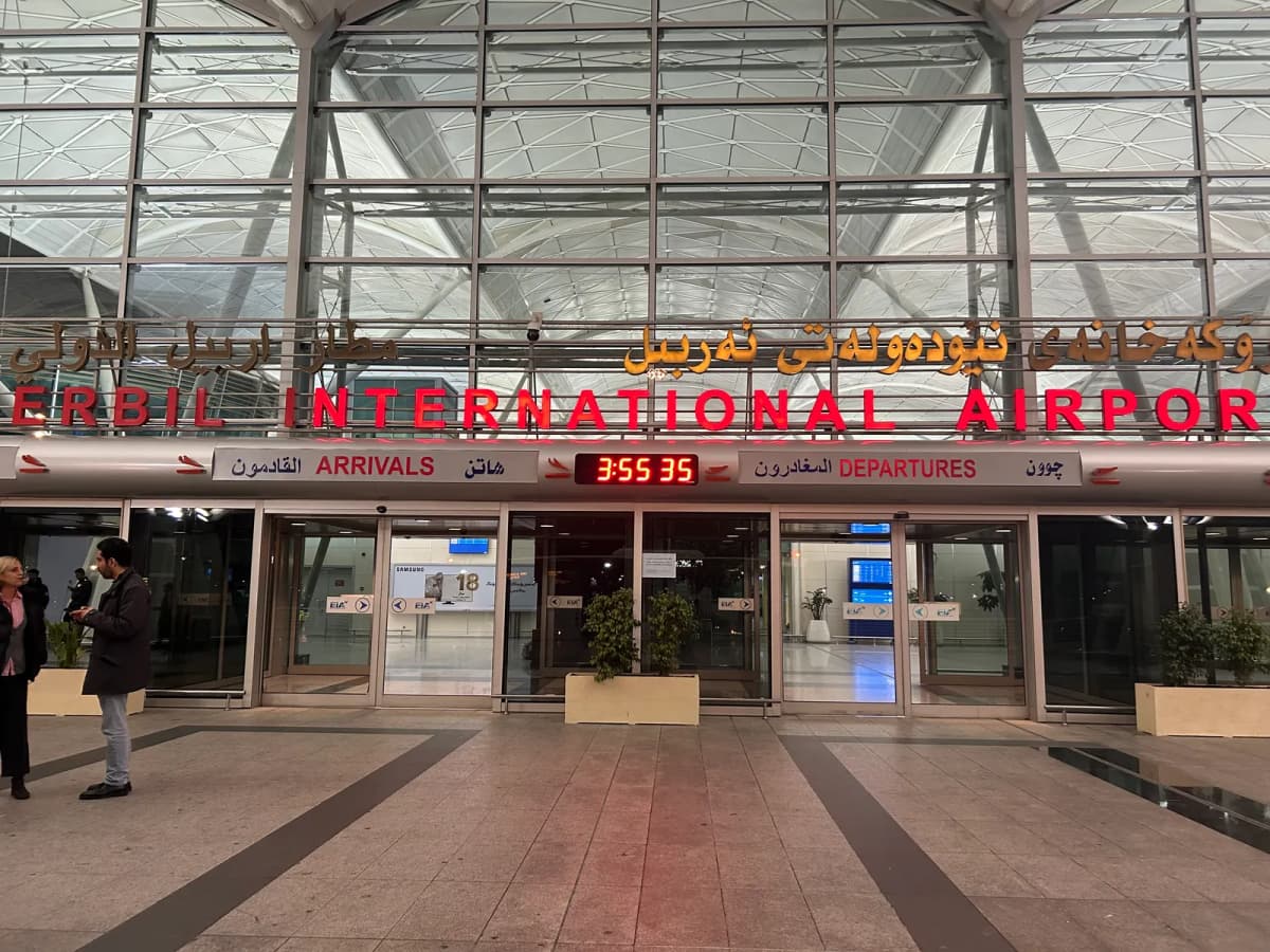 Façade vitrée d’un aéroport international, enseignes en arabe, kurde et anglais au-dessus des portes.